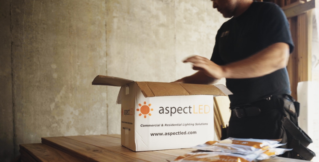 An electrician unpacking aspectLED lighting from a branded cardboard box on a wooden workbench.