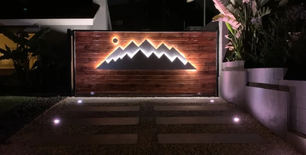 Wooden outdoor entrance sign featuring a backlit mountain design with warm LED lighting, surrounded by stone landscaping, stepping stones, and decorative plants at night.