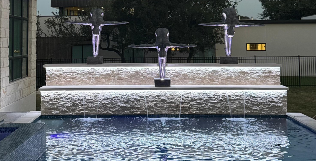 Backyard pool with a stone waterfall, illuminated water features, and three statues at night