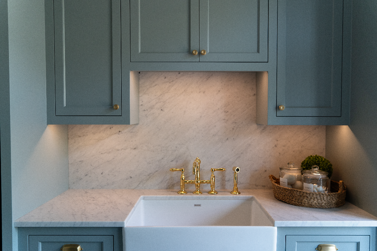 Green cabinets with task lighting above a sink with gold fixtures and a basket with supplies on the counter 