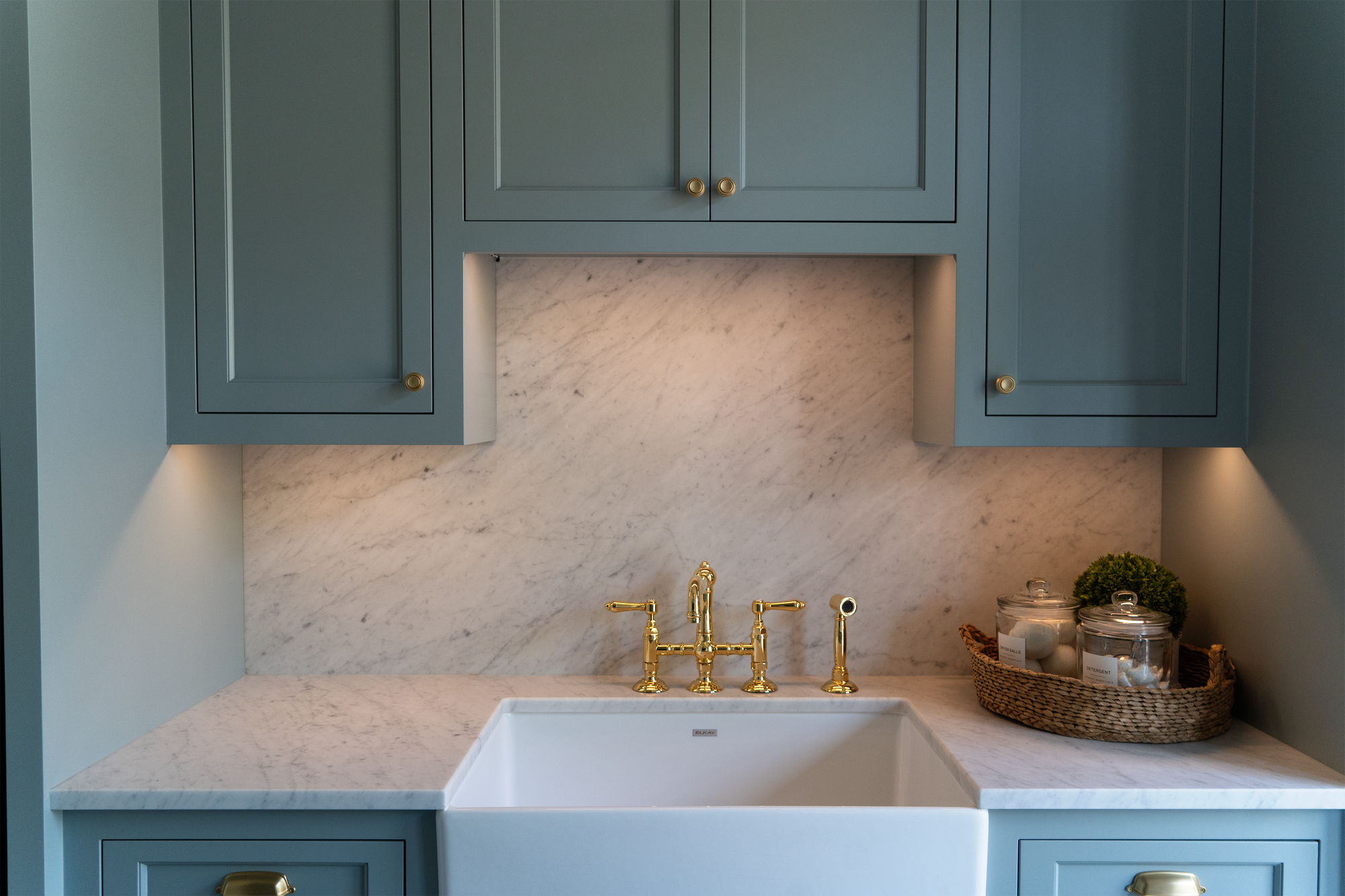 Green cabinets with task lighting above a sink with gold fixtures and a basket with supplies on the counter 